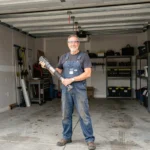 Un homme en salopette tenant un vérin de porte de garage devant une porte ouverte, dans un garage résidentiel bien rangé